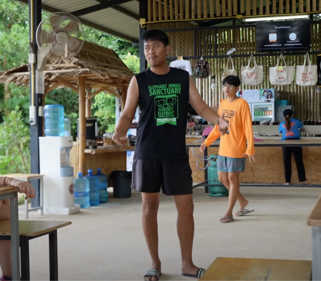 Scène montrant l'arrivée au Elephant Jungle Sanctuary Phuket, avec un éléphant et un guide local accueillant les visiteurs près d'un panneau du sanctuaire.